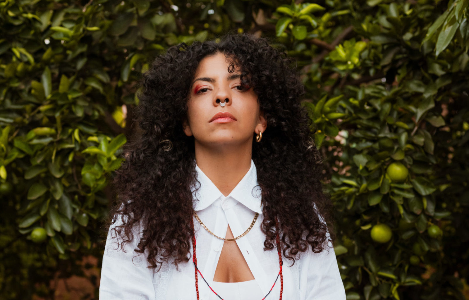 Luara Raio pose devant un pommier, la tête haute et l’air sérieux. Luara a de longs cheveux bouclés bruns foncés, coupés en dégradé, un piercing doré au septum, un boucle d’oreille à l’oreille droite, dorée aussi. Luara porte une ombre à paupière rouge-rose, une chemise blanche et trois colliers: un en or, et les deux autres sont des colliers en perle.